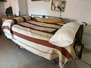 Cama articulada tipo hospital con barandillas, colchón y colcha de rayas rojas y beige. Se encuentra en una habitación individual con decoración personalizada en la pared.