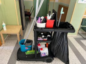 Carro de limpieza profesional con cubos, escoba, productos de limpieza, bolsas de basura y utensilios organizados. Se encuentra en el pasillo del centro.