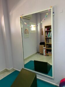 Espejo de cuerpo entero montado en la pared de una sala de terapia, decorado con una guirnalda de colores. Frente al espejo, colchonetas y material de trabajo corporal.