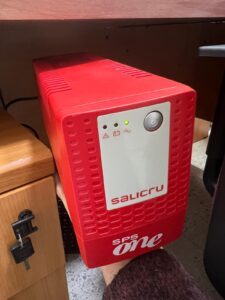 Dispositivo de alimentación ininterrumpida (SAI) de color rojo marca Salicru, modelo SPS One. Ubicado debajo de un escritorio, proporciona respaldo eléctrico en caso de cortes de luz.