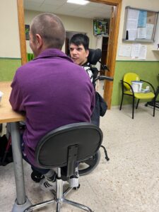 Persona en silla de ruedas participando en una conversación con una profesional, ambas en un entorno interior de atención o terapia. La escena transmite cercanía y comunicación en un entorno accesible y acogedor.