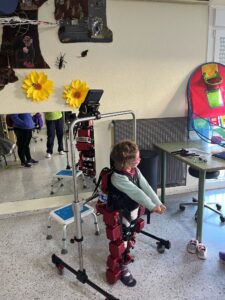 Niña con gafas y cabello rizado de pie con ayuda de un exoesqueleto de rehabilitación. La niña extiende los brazos mientras el dispositivo la sostiene en posición vertical en una sala con decoración infantil y equipo terapéutico.
