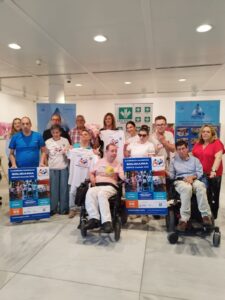 Grupo de personas, algunas en silla de ruedas, posan sonrientes con camisetas y carteles promocionales de la 10ª Carrera-Marcha Solidaria ASPACE Granada 2025. Se observa diversidad de edades y capacidades, mostrando un ambiente inclusivo y alegre.
