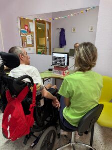 Una profesional señala la pantalla de un comunicador digital mientras acompaña a un hombre en silla de ruedas. Ambos sonríen y comparten un momento de interacción en un entorno cálido, con decoración colorida y estanterías con materiales didácticos