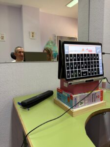 Primer plano de un dispositivo con un teclado virtual adaptado, apoyado sobre varias cajas para elevarlo a la altura adecuada. En la pantalla se muestra una frase escrita. Al fondo, un hombre sonríe mientras se refleja en un espejo junto a la profesional que lo acompaña.