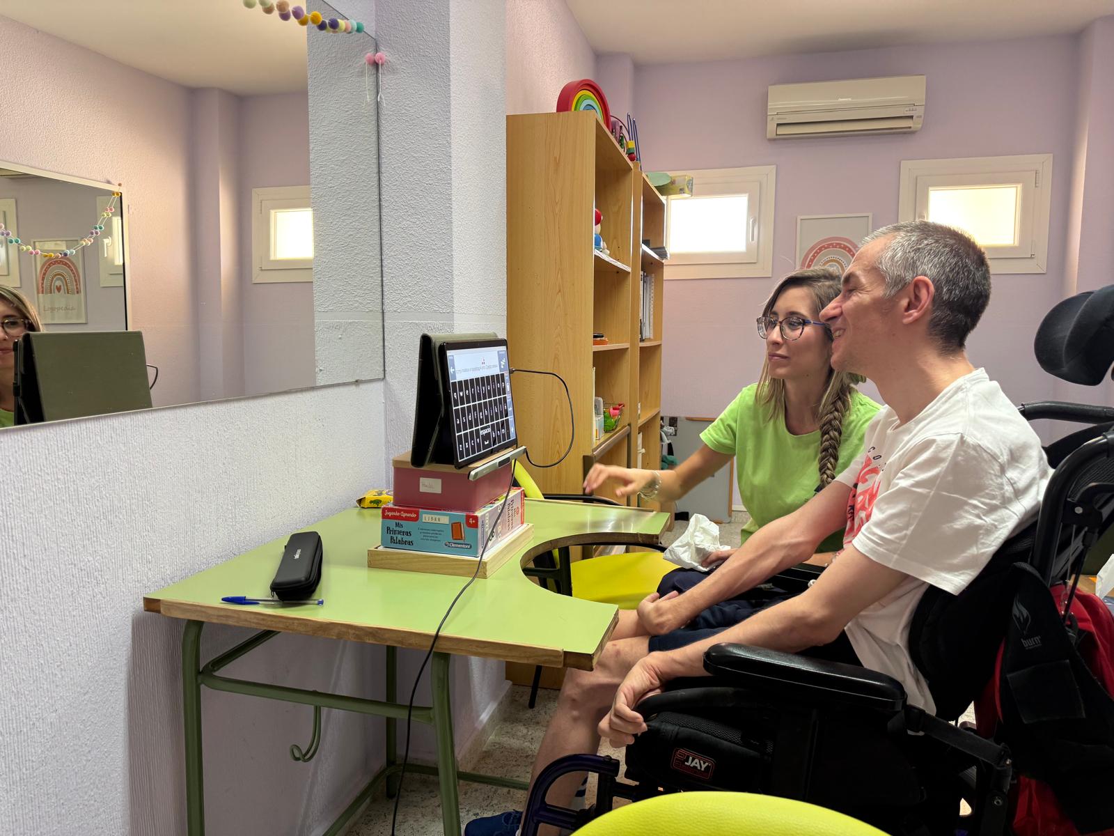 Un hombre en silla de ruedas con una mochila roja y una profesional sentada junto a él utilizan un sistema de comunicación aumentativa con un teclado en pantalla. Ambos están sentados frente a una mesa en una sala decorada con colores suaves y elementos infantiles.