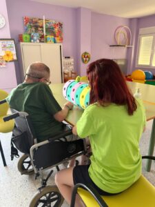 Una persona en silla de ruedas y una profesional participan en una actividad de estimulación sensorial en una sala ocupacional. Ambos están sentados frente a una mesa y manipulan un cilindro blando de colores vivos con textura y formas, diseñado para trabajar la coordinación y la atención. El entorno es luminoso, con paredes lilas decoradas con juegos, dibujos y material lúdico-terapéutico. La escena refleja un ambiente inclusivo, dinámico y adaptado a las necesidades de personas con discapacidad.