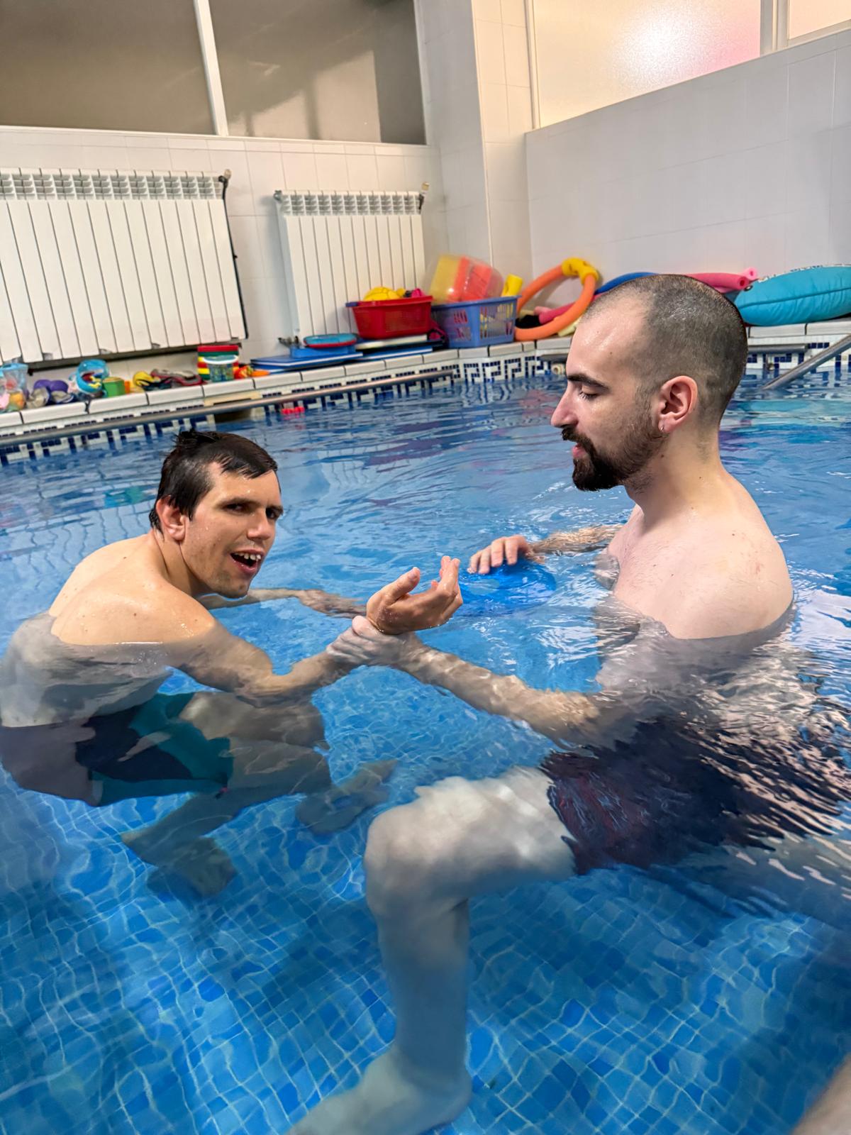 HABILITACION 5 Dos personas adultas participan en una sesión de habilitación acuática en una piscina interior. Una de ellas, con parálisis cerebral, es guiada por un terapeuta que sostiene su brazo mientras ambos están parcialmente sumergidos. La actividad se desarrolla en un entorno adaptado, con materiales de flotación y seguridad visibles al fondo. La escena muestra una intervención terapéutica enfocada en la movilidad, el equilibrio y el desarrollo sensoriomotor en un medio acuático.