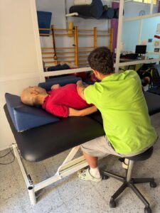 Un profesional realiza una sesión de habilitación física con una persona adulta con parálisis cerebral, que está tumbada de lado sobre una camilla de fisioterapia. La intervención se lleva a cabo en una sala equipada con material terapéutico como espalderas, rulos y cojines posturales. La escena refleja un trabajo individualizado orientado a mejorar la movilidad y el bienestar corporal de la persona atendida.