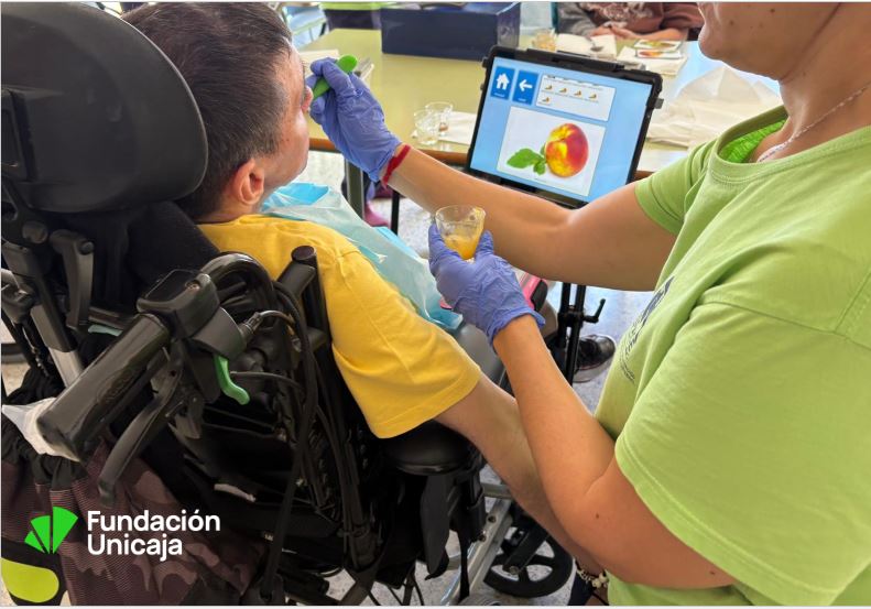 Persona con parálisis cerebral en silla de ruedas recibe comida texturizada de una profesional, mientras en una tablet se muestran pictogramas de alimentos. Logo de Fundación Unicaja en la esquina.
